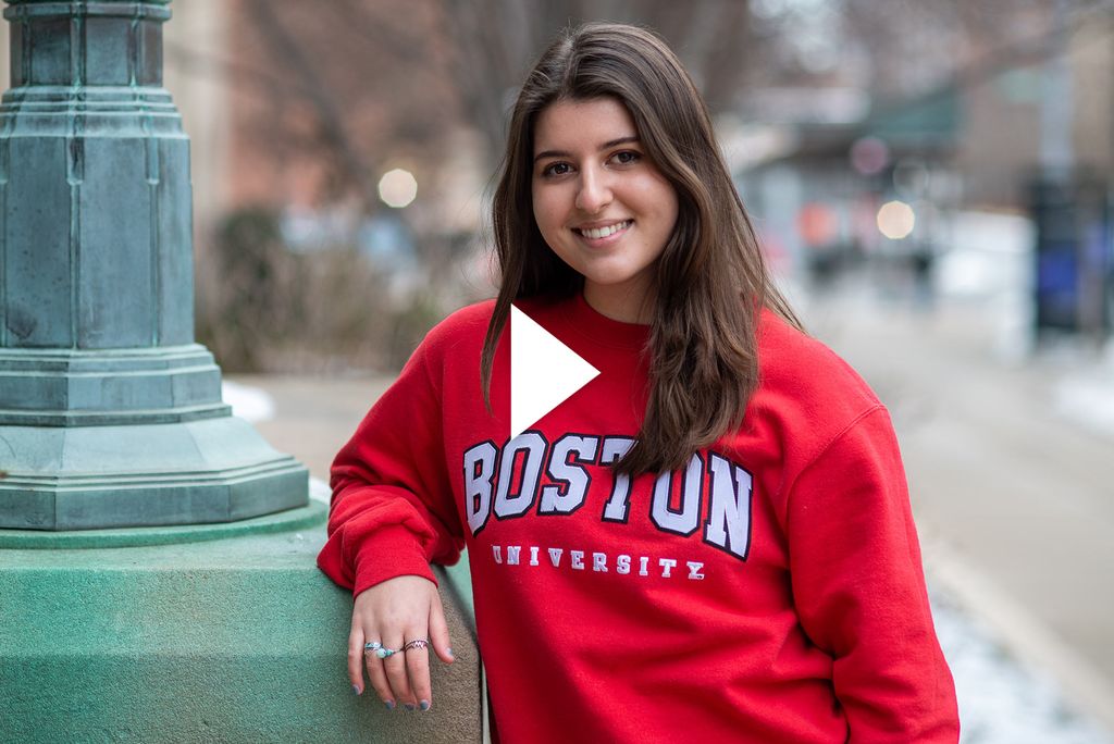 Photo of Vlogger Anna Malygin (Questrom'23), smiling, with her hand resting on the base of a green lamp post. She wears a bright red Boston University Sweatshirt, and has her hair down. Video play button is overlaid.