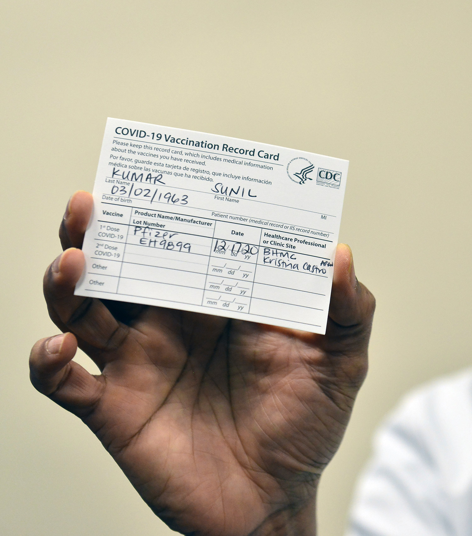 Photo of someone holding up their vaccination card. It reads “COVID-19 Vaccination Record Card….Last Name Kumar, First Name Sunil,” and other personal information. They received the Pfizer vaccine. The background is beige.