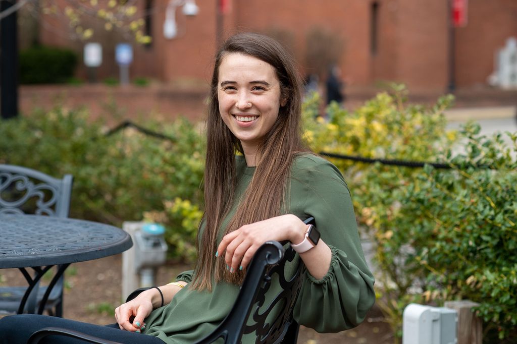 Student Employee of the Year Manisha Cole, sitting on a metal chair outside, and smiling. She has long brown hair and wears a green blouse.