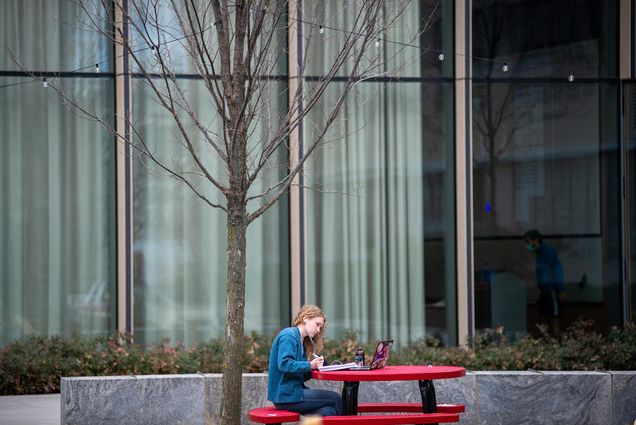 Photo of Anna Scholz-Carlson (COM’21) sitting a red picnic table on the COM Lawn looking at her laptop as she attends class virtually. She wears a blue jacket.