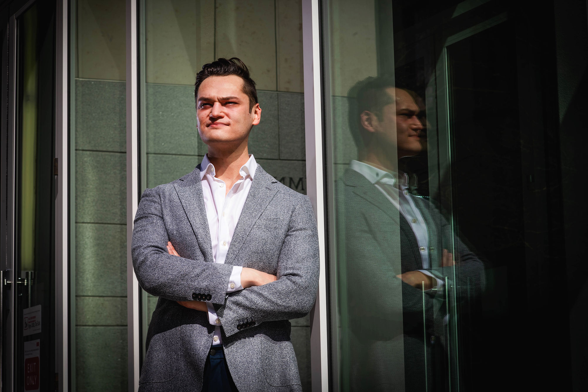 Photo of Eduardo Gonzales (LAW’21), in a gray, speckled suit jacket, with his arms crossed, he looks determinedly off into into the middle distance. He leans against the windows of a large building.