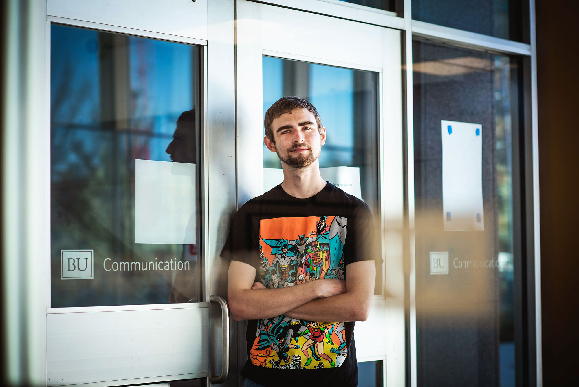 Photo of Richard Boylan (COM'22) in a black marvel super heroes shirt with his arms crossed, slightly smiling, as he leans against the entrance of one of the COM buildings.