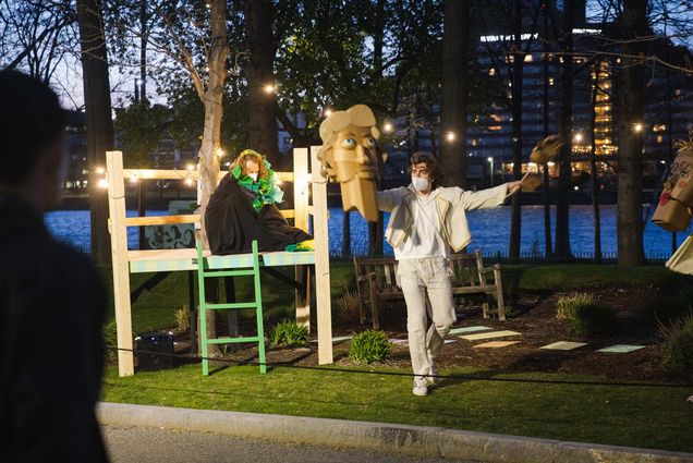 Photo of Michael Ticknor (Demetrius), right, and Matthew Swain (Oberon), left, perform during the pop-up outdoor production of A Mid-Summer Night’s Dream, directed by Chaal Aydiner on Thursday, April 22nd. Swain wears all white and beige clothes and holds up a wooden head. Ticknor sits on top of a platform, wrapped in a black blanket and green ruffles. They perform along the Charles River esplanade.