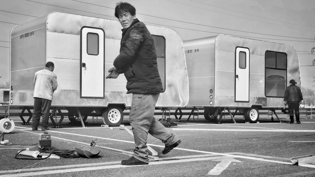 Black and white photo of a man as he turns towards the camera; behind him are small trailers, and he stands in what looks like a parking lot.