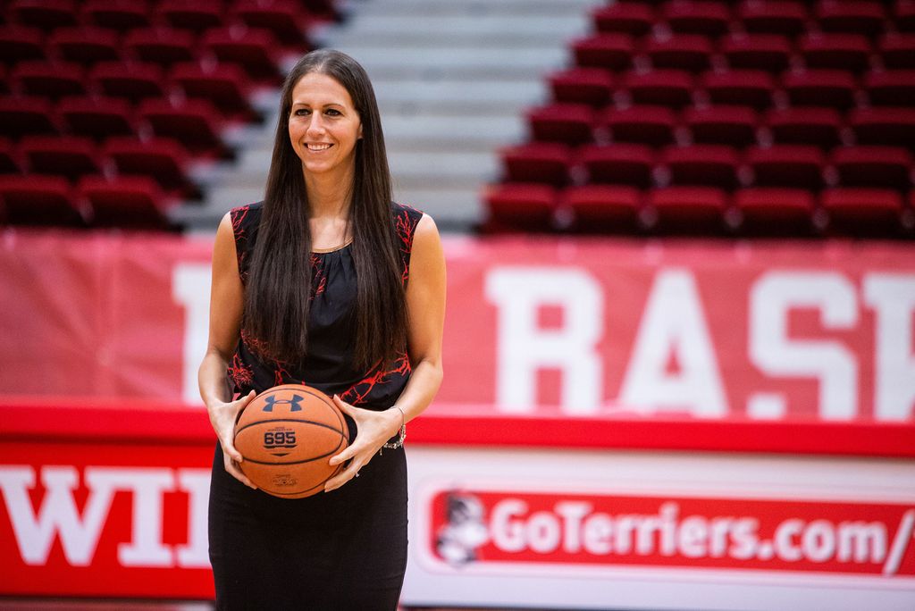Photo of new women’s basketball head coach Melissa D’Amico, standing and holding a basketball, she wears  a red and maroon textured blouse, and has long brown hair. A sign behind her in the gym reads “GoTerriers.com/…”