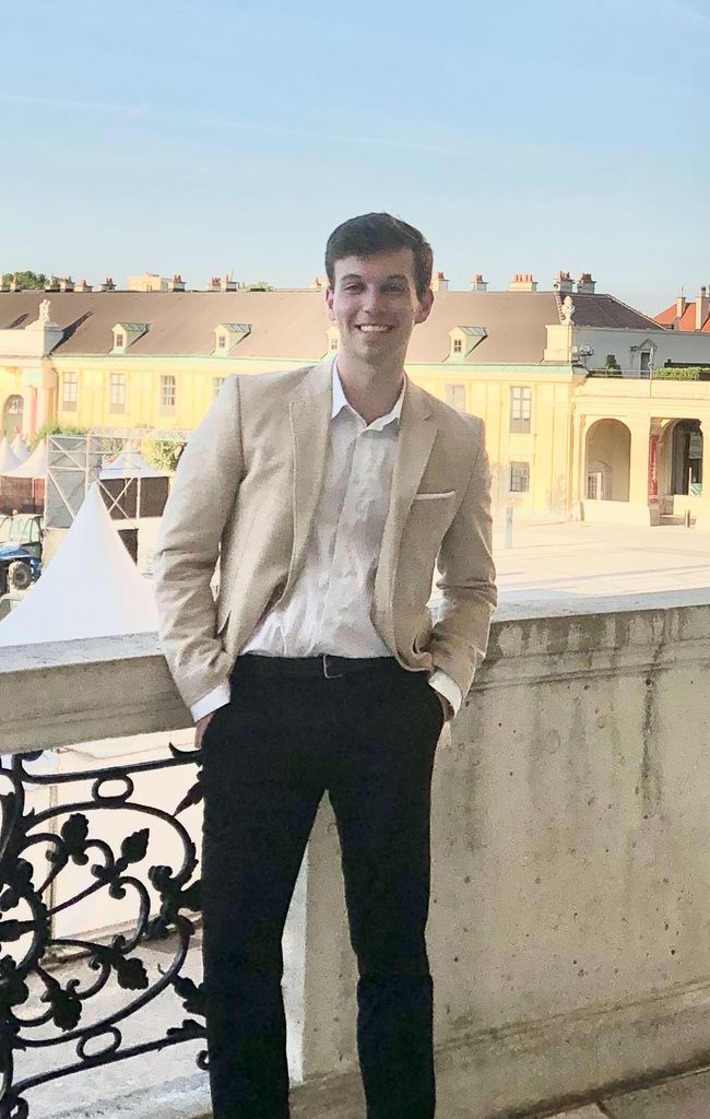 Photo of Eliot Usherenko (Pardee’21)in a tan blazer and black pants, smiling, with his hands in his pockets, on a balcony, with a large piazza in the background.