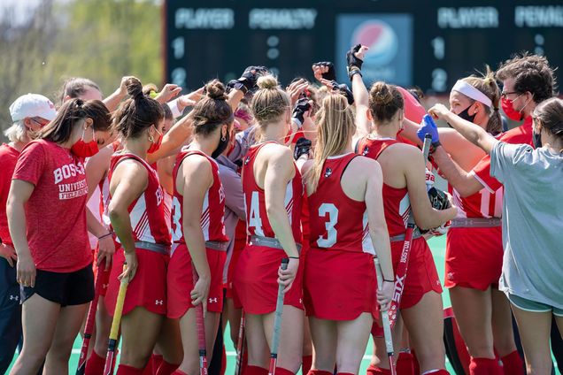 Photo of the BU field hockey team in a huddle; the women wear red jerseys and shorts and hold their sticks, some coaches are seen as well. They all wear face masks and raise their hands towards the center.