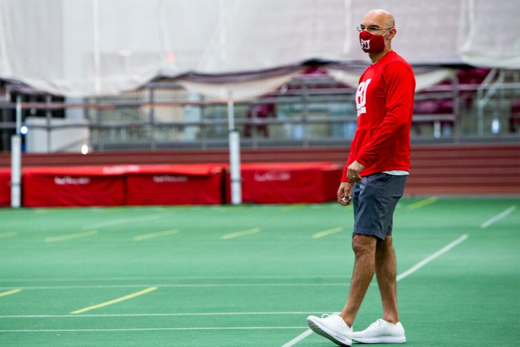 Photo of Gabe Sanders, BU’s director of track and field, standing in the middle of the indoor track, which has green flooring. He wears a red, BU long sleeve, and wears gray shorts.