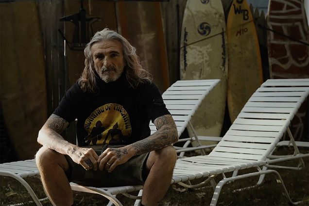 Photo of a man with long gray hair sitting on a white plastic reclining lawn chair with his elbows resting on his news. He wears shorts and a black and gold t-shirt and looks honest and content.