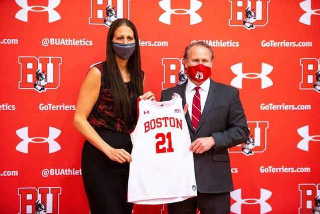 Photo of Boston University Director of Athletics Drew Marrochello (right), in a red BU face mask and suit, presents new women’s basketball head coach Melissa D’Amico, left, in a red and maroon textured blouse, with a jersey with the number 21 during D’Amico’s official introduction during a virtual press conference on Monday afternoon. The two stand before a red backdrop with the BU athletics logo on it.