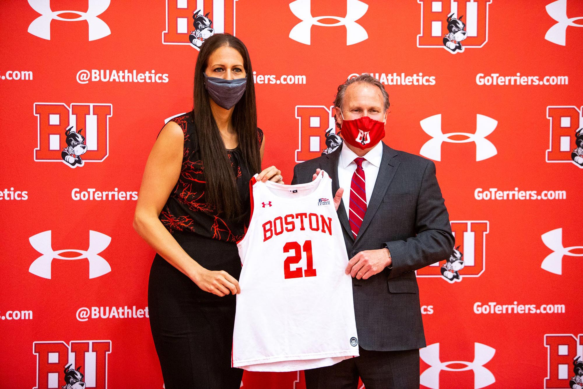 Photo of Boston University Director of Athletics Drew Marrochello (right), in a red BU face mask and suit, presents new women’s basketball head coach Melissa D’Amico, left, in a red and maroon textured blouse, with a jersey with the number 21 during  D’Amico’s official introduction during a virtual press conference on Monday afternoon. The two stand before a red backdrop with the BU athletics logo on it.