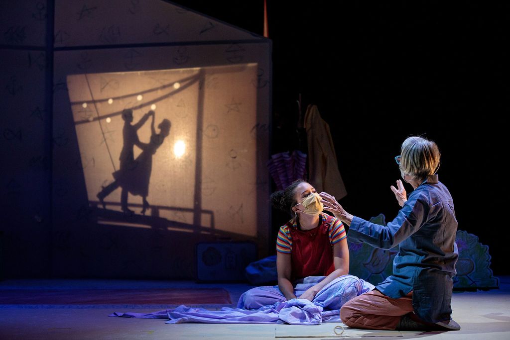 Photo of Elaine Vaan Hogue (CFA’97), left, and Valyn Turner (CFA’23) play Grammy Sophie and her granddaughter Esme in the Wheelock Family Theatre virtual production of Walking the Tightrope. The pair sits on the ground, Hogue extends her hands as if telling a story and Turner looks at Hogue engrossed in the story. In the background, two shadow puppets dance on the wall.