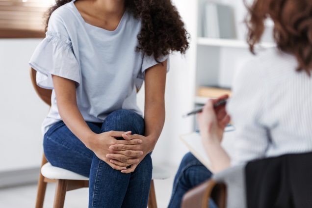 Photo of a young person in jeans and a light blue shirt with their hands over their crossed legs, sitting on a chair talking to a therapist or counselor of some kind, that is seen blurred on the right foreground.