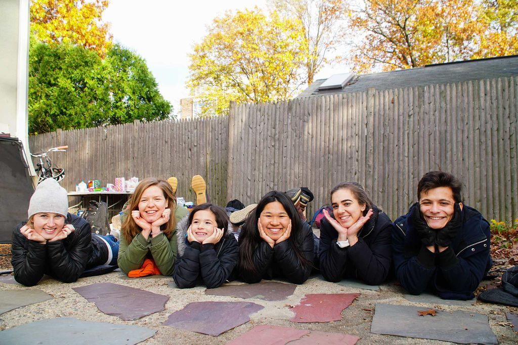 A photo of several young people laying on the ground with their faces in the their hands. Behind them is wooden fence and trees with fall leaves.