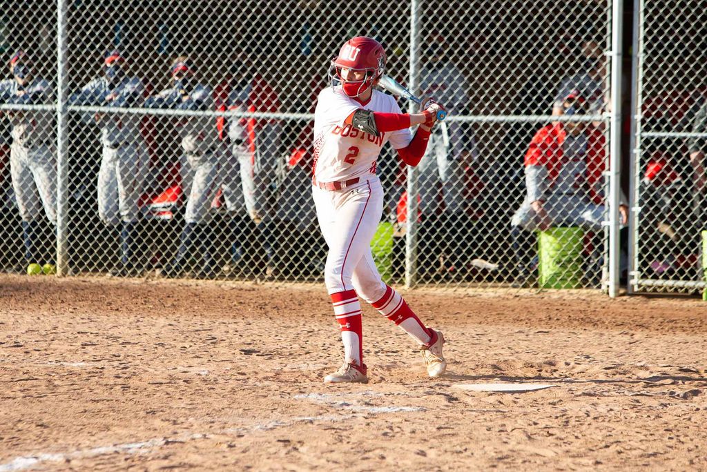 Photo of the women’s softball player Emily Gant, number 2, mid-swing during her turn at bat. Behind her, her teammates lean on the fence and watch the player.