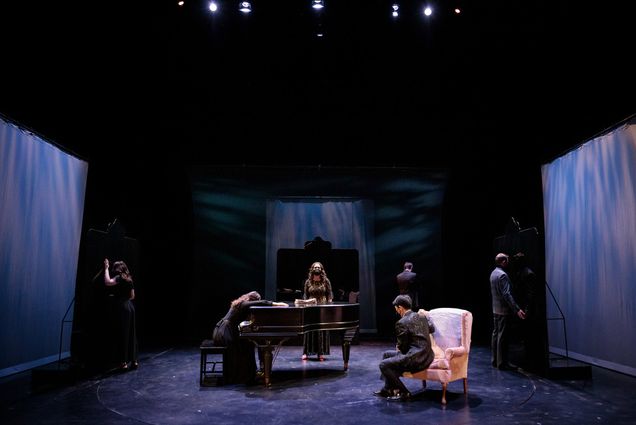 Photo of the cast of Mansfield Park on stage, presumably during a dress rehearsal. At center, a woman in a black dress drapes herself over a grand piano. A man sits in a fancy pink chair. At the edges of the stage, people stand with their faces away from the camera. Behind the piano, a woman in a golden dress and mask stands.