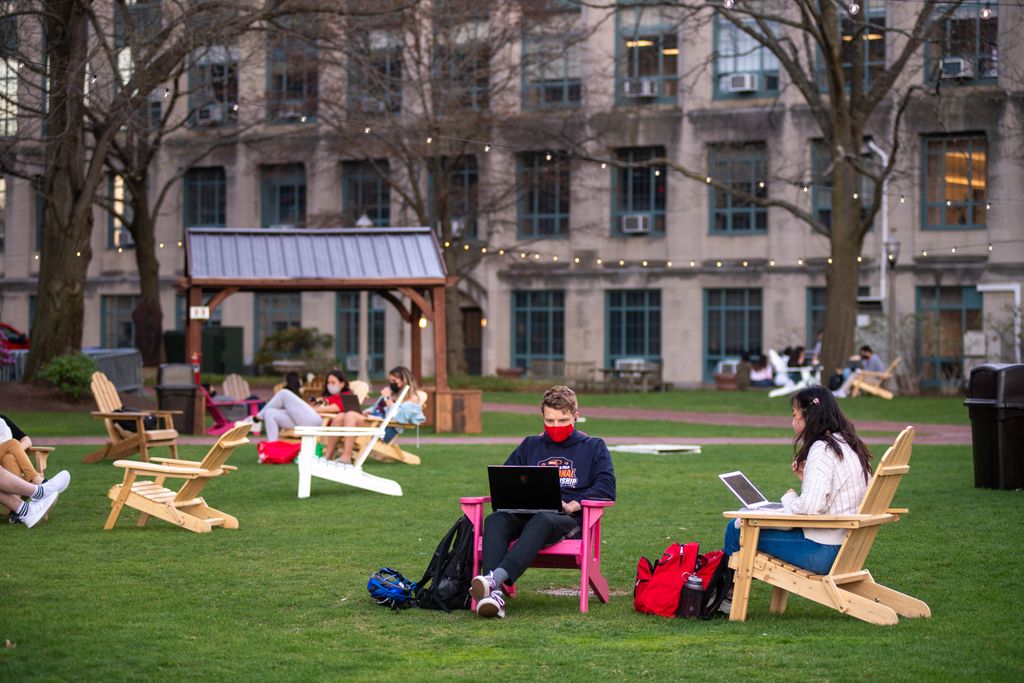 Photo of Julia Lee (SAR’22), right, and Jack Gardiner (SAR’22) sitting under the lights on BU Beach in Adirondack chairs and working on their laptops.