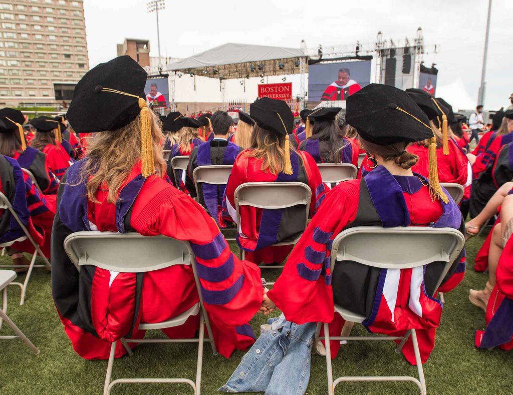 View of the stage and screen from behind seated students at the 2021 Commencement cermony for advanced degrees. Noubar Afeyan can be seen on the screens delivering his address.