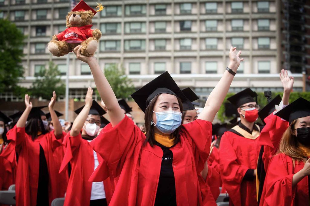 A group of graduates hold their arms in the air and cheer during Commencement. The woman at the fron of the group is holiding a BU Graduate teddy bear with red mortarboard.