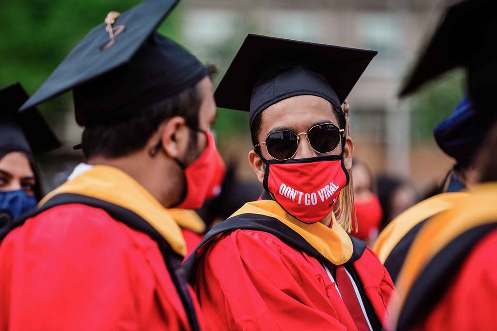 A graduate wearing black mortarboard, red robe with black and gold stole, sunglasses and a PPE face covering that says 'Don't Go Viral' talks to another graduate on Nickerson Field.