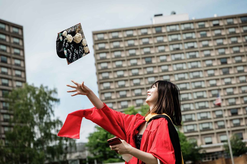 A graduate tosses her decorated mortarboard cap in the air on Nickerson Field during BU's 2021 Commencement. BU dorm buildings can be seen in the background.