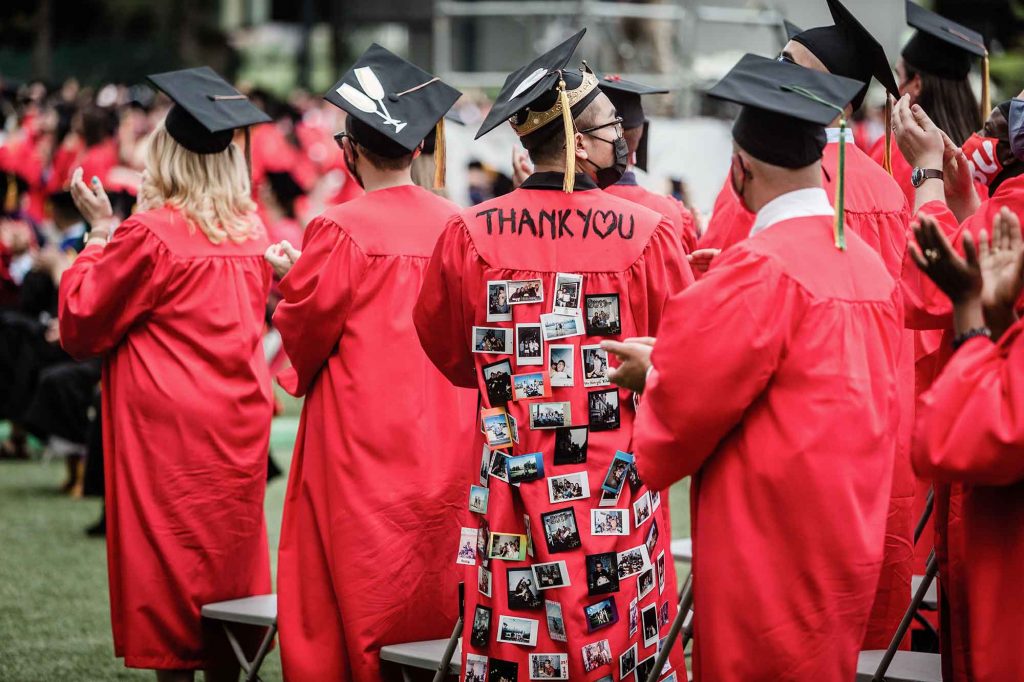 Back view of a student whose robe says 'THANK YOU' (the O written as a heart) with many photos pinned to the back of the robe.