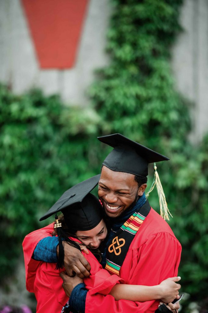 2 graduating students wearing cap and gown embrace in a big hug following Commencement