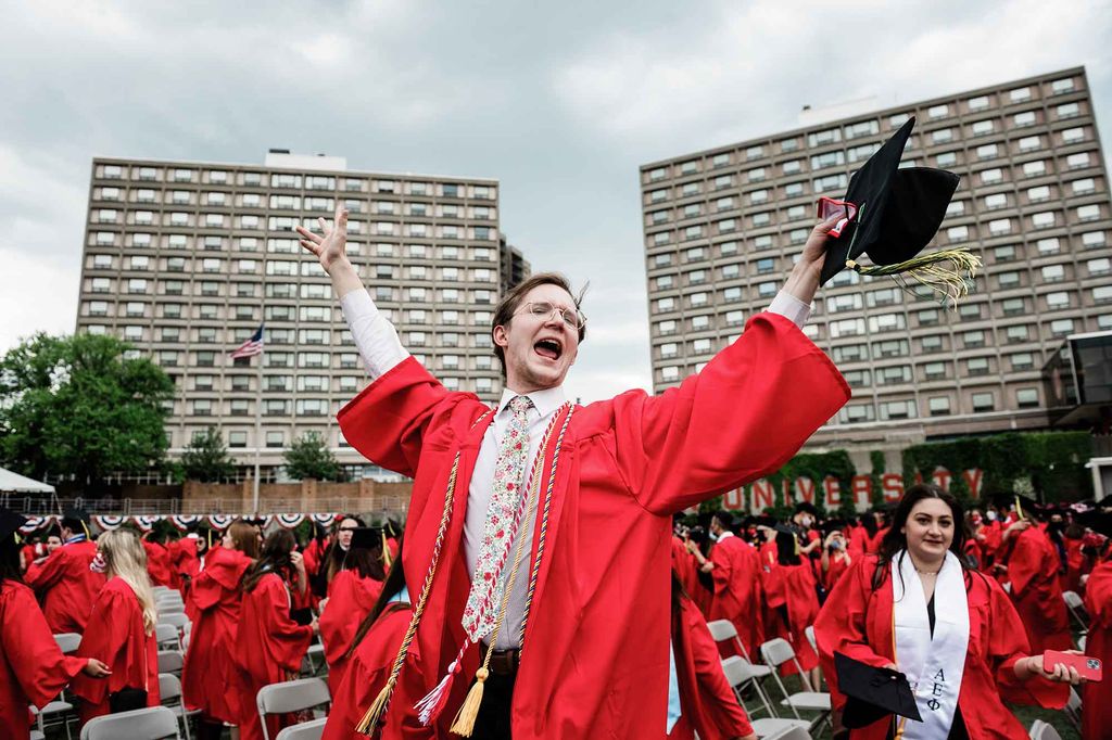 A graduate wearing red robe with colored honor cords holds his hands in the air and cheers.