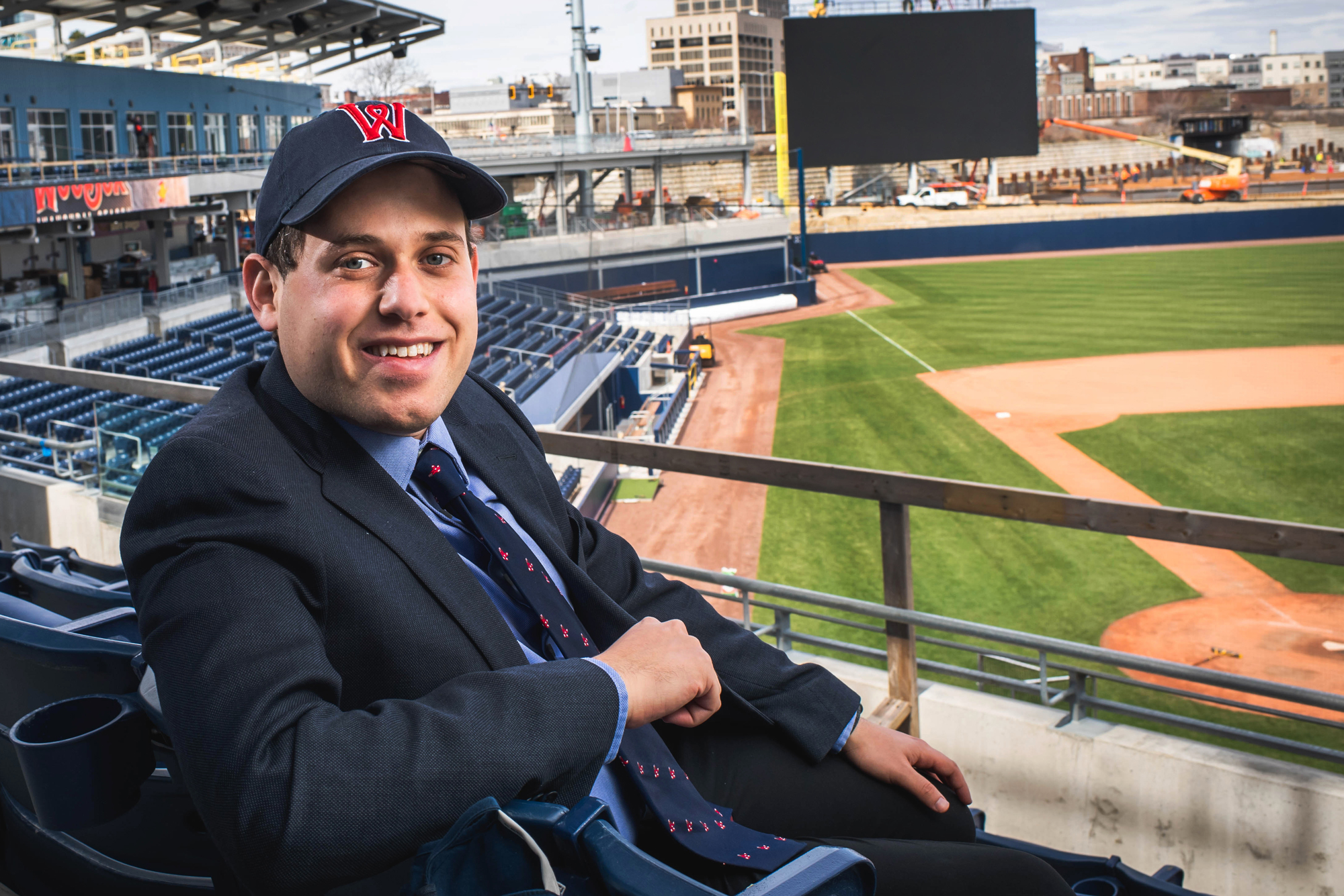 A photo of Matt Levin wearing a Worcester Red Sox hat sitting in the stands. The baseball field is in the background.