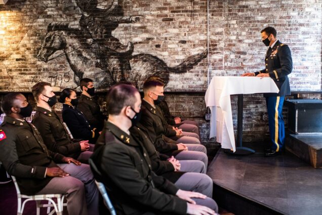 Photo of LTC Nathan Luecke speakubg during the ROTC Commissioning ceremony at Black Rose Tavern on Monday, May 17, 2021. The photo is taken from his left side and shows him in profile speaking at a table covered with a white tablecloth two a few rows of cadets seated with black face masks on.