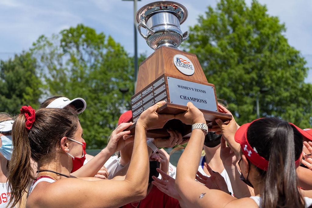 Photo of the BU women’s tennis team hoisting up the trophy as a group after winning the championship match. The women wear red bows and visors and have face masks on. The trophy is silver and sits in a wooden base with plaques on it.