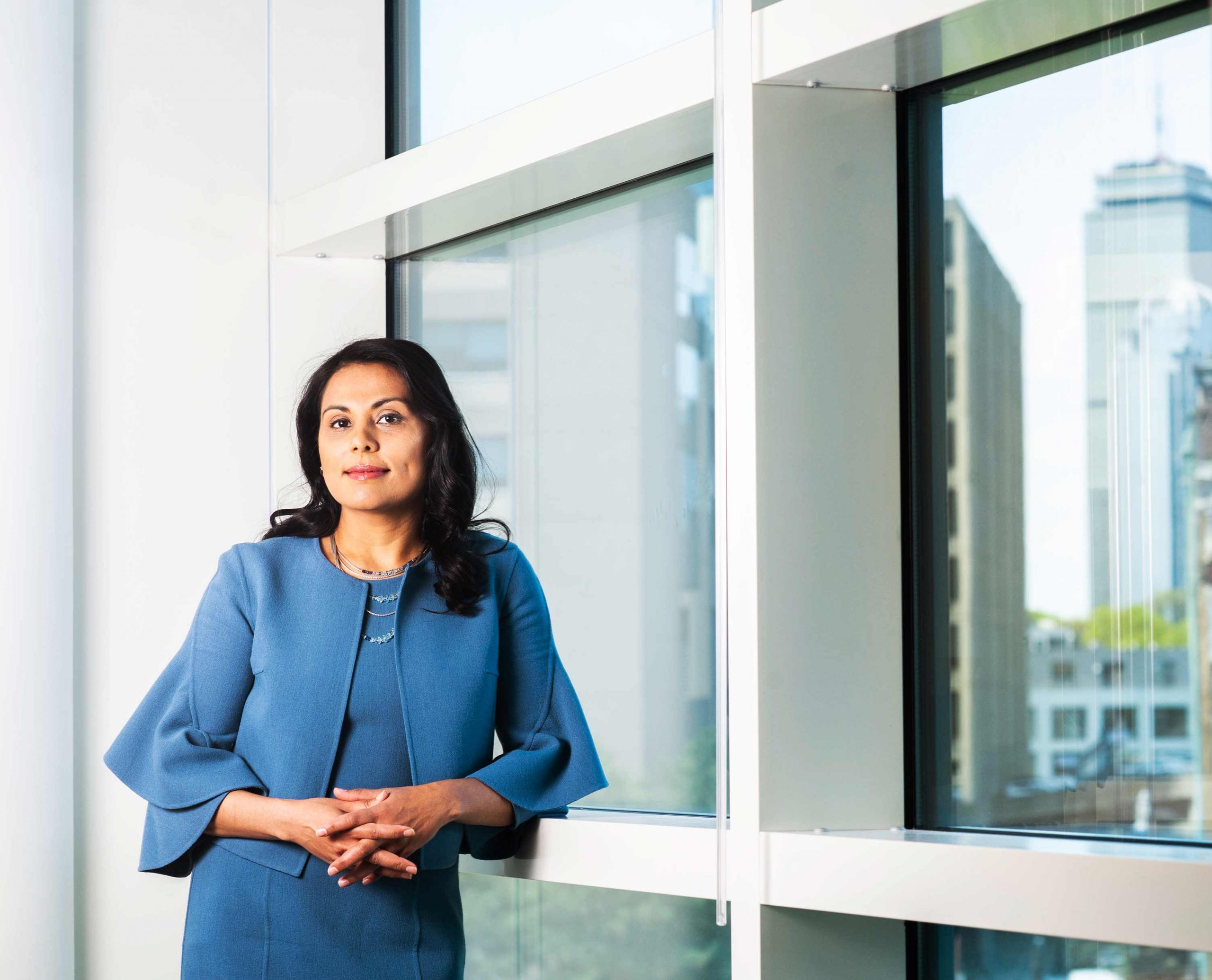 Portrait of Dr. Nahid Bhadelia leaning against a building in front of a window showing a reflection of the Boston skyline.