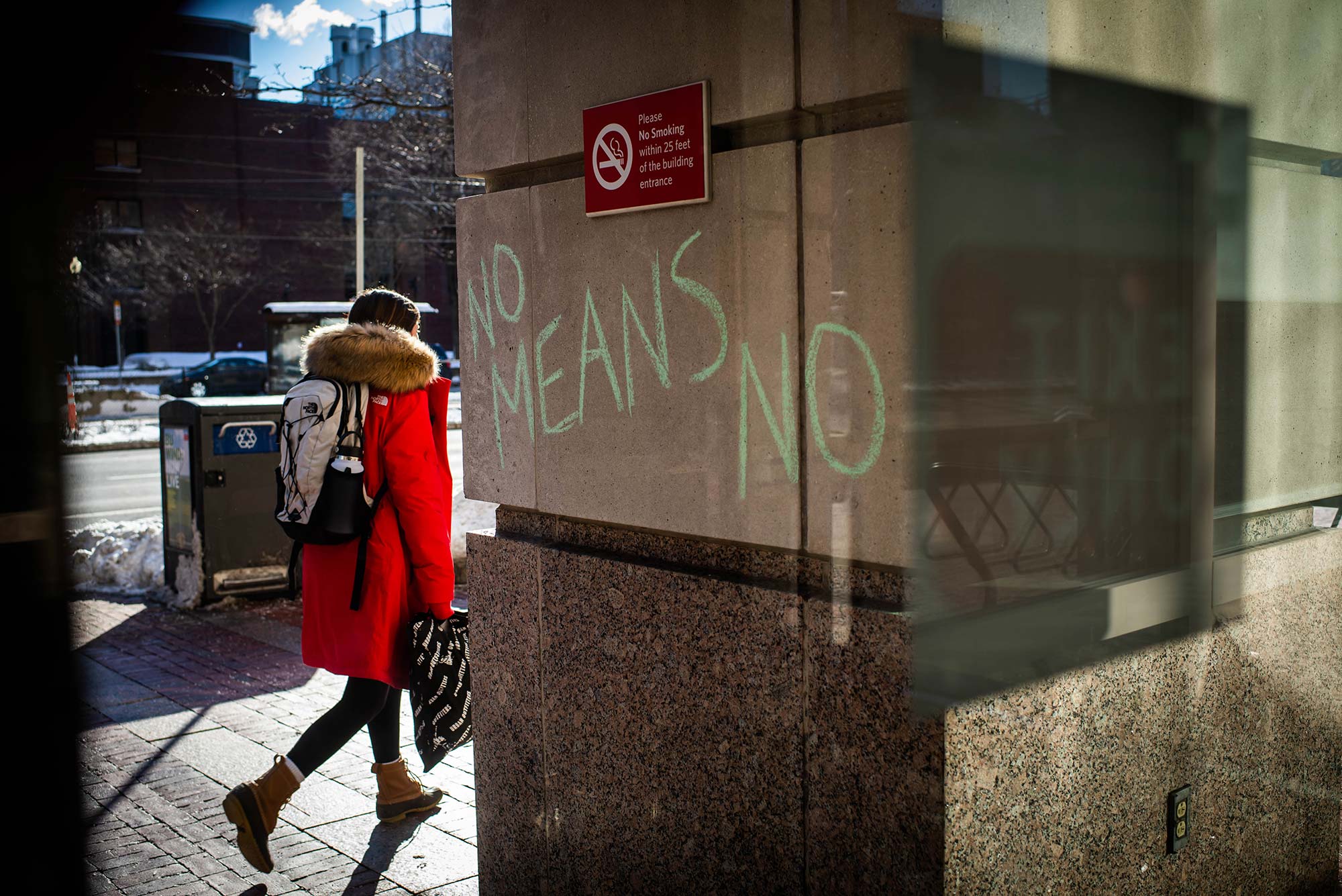 Photo of a cement building column that has the words "No means no" written in green chalk on it. A no smoking sign hangs above. In the background, a person in a red winter jacket, backpack, and bag in their hand walks down the sidewalk.
