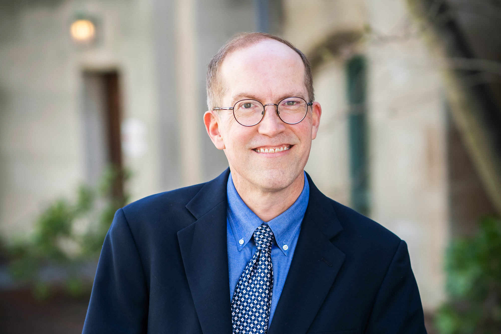 Headshot of Metcalf Award winner David Sullivan, a CAS master lecturer in computer science. He wears a blue suit and tie and has rounded glasses; he smiles as he leans on a half-wall outside.