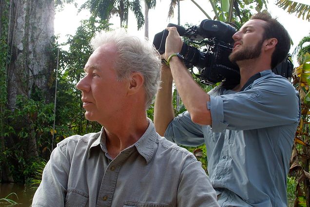 Photo of Martin Doblmeier, in profile, in a gray button up shirt looking towards his left, as a camera man behind him shoots something off in the distance. Behind them, a jungle is seen.