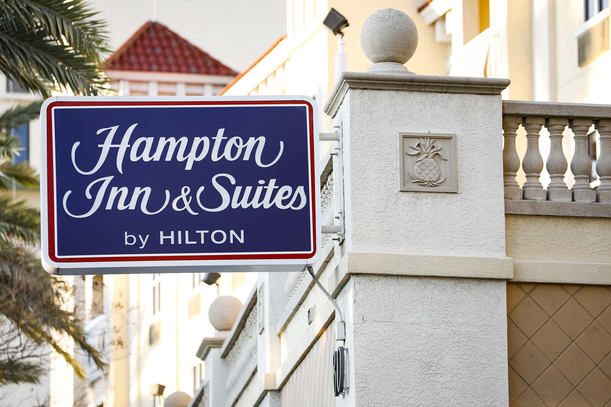 Photo of a blue “Hampton Inn & Suites by Hilton” sign hanging off the side of a stucco building with a pineapple tile at the corner. In the background, a palm tree and red roof is seen.