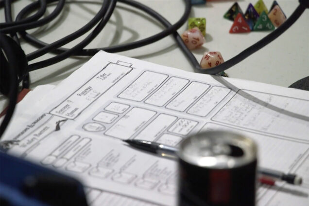 Image of a character sheet, die, mechanical pencil and microphone cords during a recording of the YMBWHWGH podcast. A blurred energy drink can is in the foreground on the bottom right.