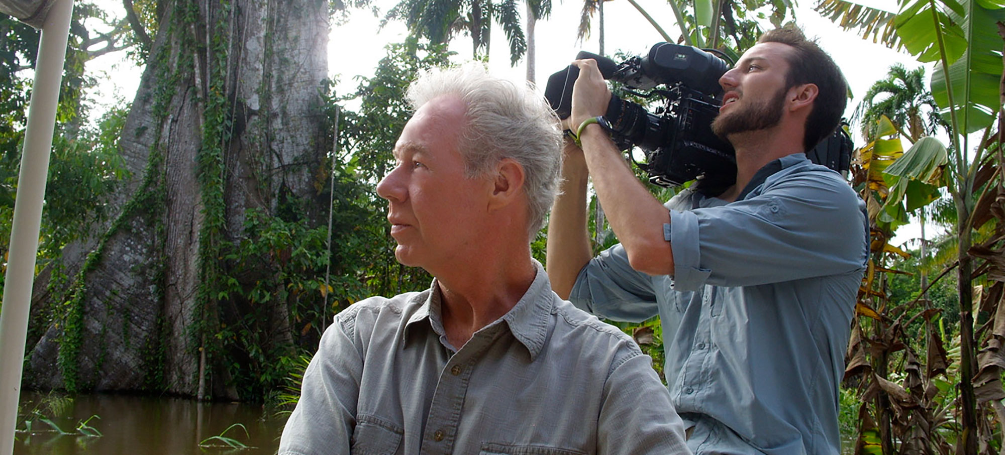 Photo of Martin Doblmeier, in profile, in a gray button up shirt looking towards his left, as a camera man behind him shoots something off in the distance. Behind them, a jungle is seen.