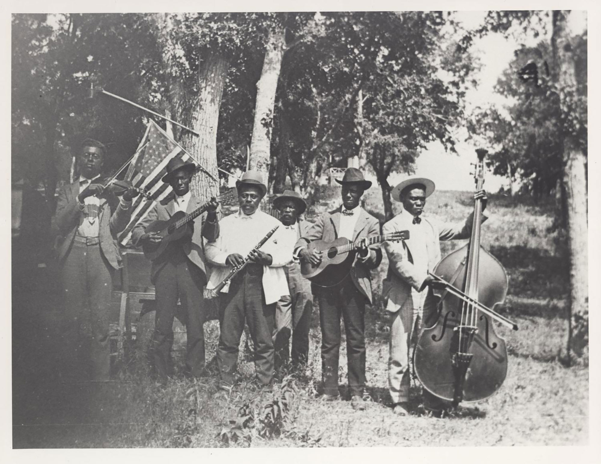 Black and white photograph of a band of African-American playing at Emancipation Day celebration in June 19, 1900, held in “East Woods” on East 24th Street in Austin. There are 6 men in the band, they all wear suits and hats and hold their instruments, including a fiddle, guitar, clarinet, and upright bass. There are trees and an American flag hanging behind them.