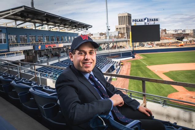 A photo of Matt Levin wearing a Worcester Red Sox hat sitting in the stands. The baseball field is in the background.