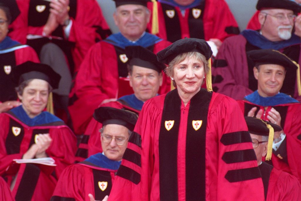 Photo of Olympia Dukakis receiving an Honorary Degree from Boston University in 2000. She wears a red gown and a black hat and smiles while looking off to her left.