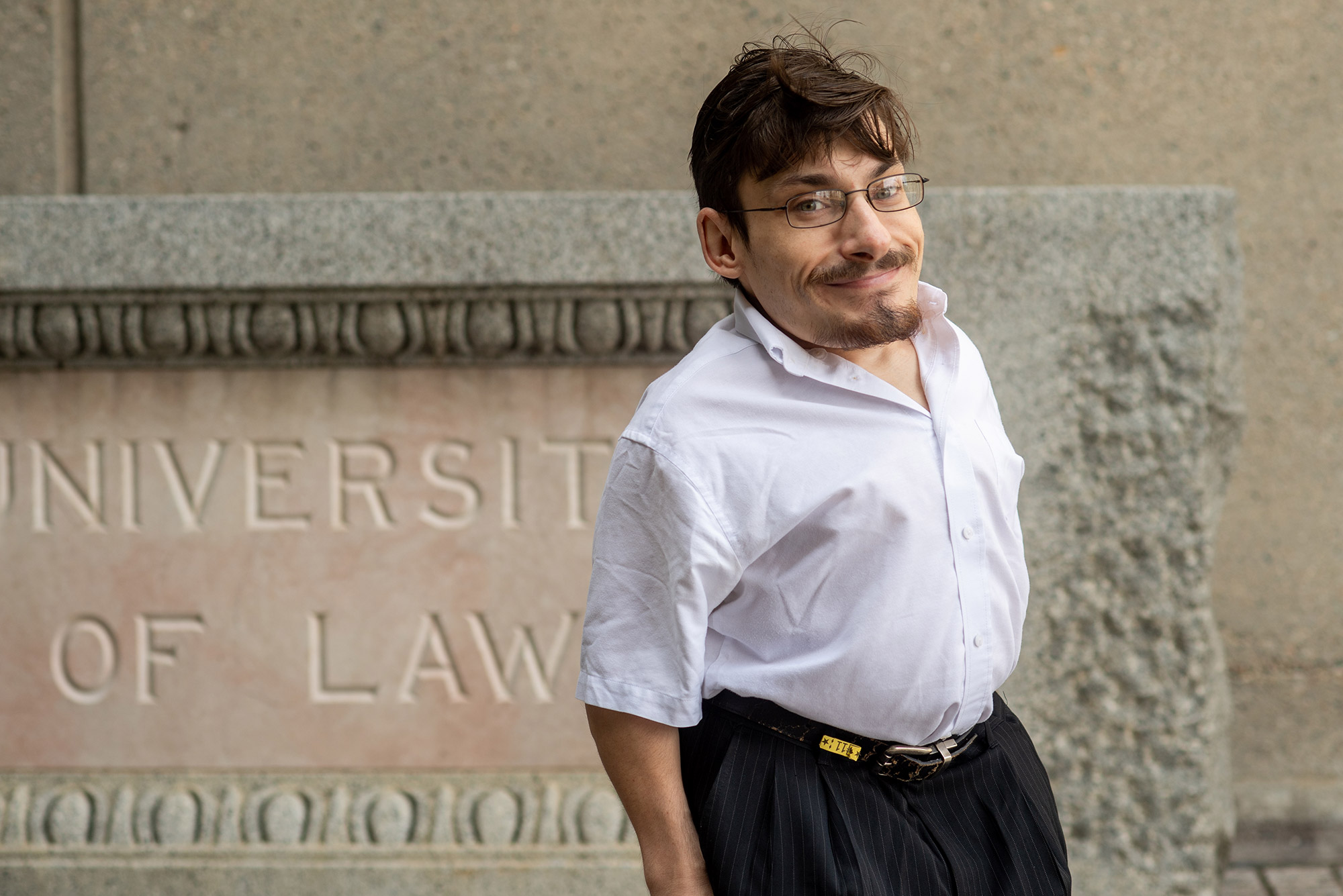 Photo of Gaetano Mortillaro in a white button down, black pants and glasses. He smiles, standing outside one of the LAW buildings and a sign “University Law” is seen in the background.