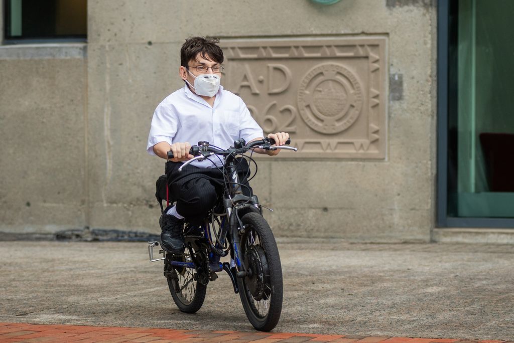 Photo of Gaetano Mortillaro in a white button down, black pants and glasses riding his black and blue bike with a face mask on. 