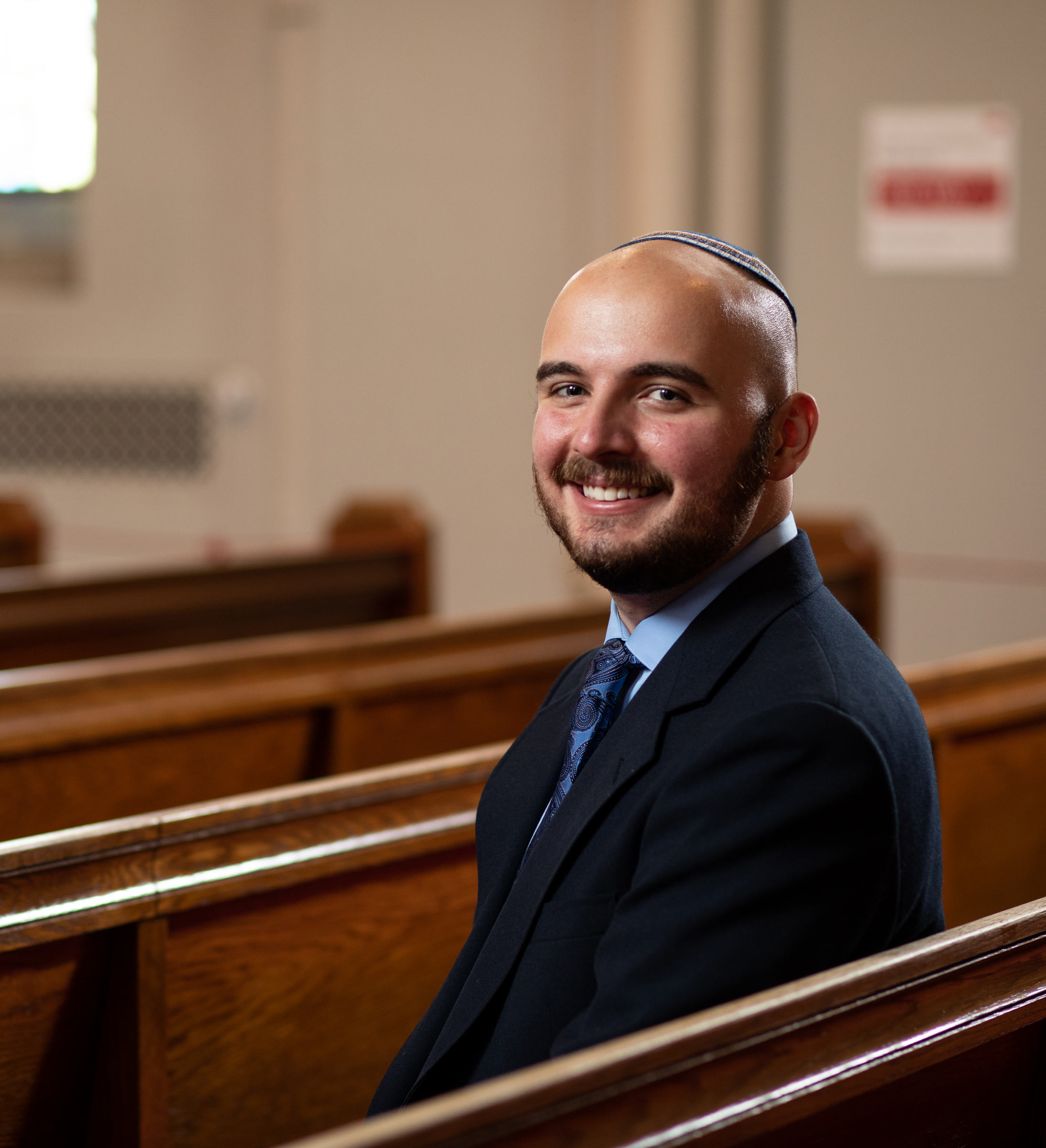 Portrait of Connor Dedrick sitting in the pews of Marsh Chapel in a suit and tie. He smiles and wears Kippah/Yamaka.