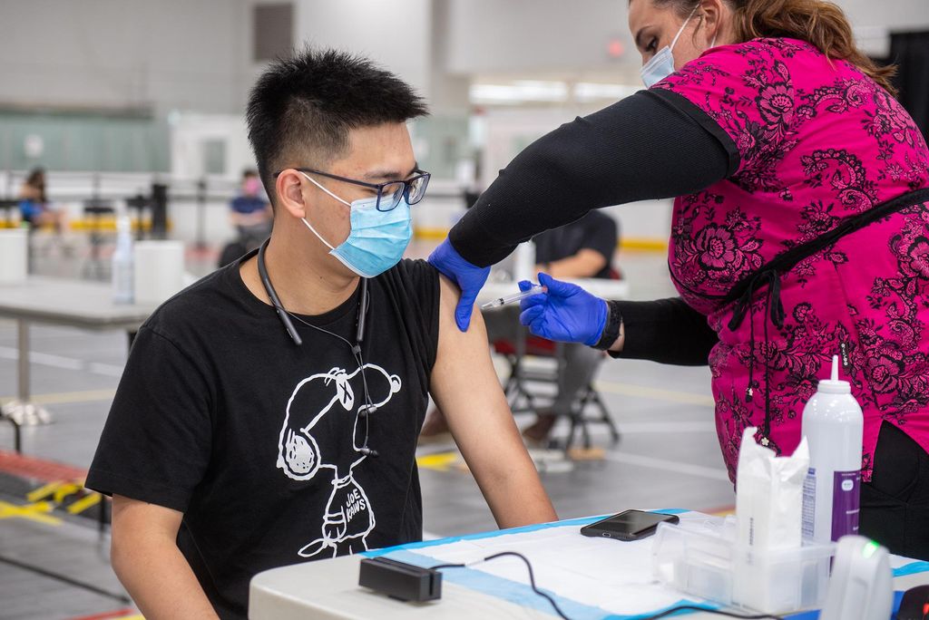 Photo of Yuqing Wu (COM’23), in a black Snoopy t-shirt, getting his vaccine from Kristin Lopes, registered nurse, in bright pink scrubs with a floral pattern, at the Moderna vaccine clinic at FitRec May 4. Both people wear masks.