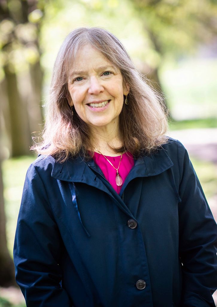 Portrait of Aline Hillman in a blue jacket and pink blouse smiling; she has shoulder length brown hair. 