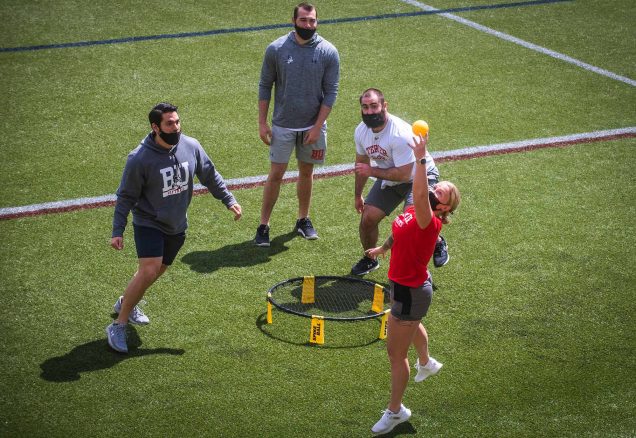 Photo of strength and conditioning coaches Massimo Gioffre, Sam Greenfield, Chris Fucillo and Claire Sporer play a game of spike ball on Nickerson field on May 13, 2021. Claire, who wears a red shirt and shorts, reaches up to spike the ball to her partner. The others wear BU athletics gear, like sweatshirts, t-shirts and shorts with the BU logos.