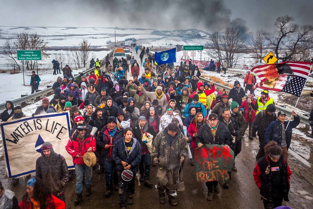 Photo of a group of indigenous water protectors and their allies marching down a road in protest of the Dakota Access Pipeline during the Standing Rock protests in North Dakota. The protestors are a mixture of old and young people, bundled up in winter gear. The hold signs that say “Honor the Treatys,” “Water is Life,” and one with an image of the earth. Another person holds a desecrated US flag. Behind them, a green highway sign on the left reads “Leaving Standing Rock Indian Reservation,” another on the right reads “Cannonball River” and folks in the background hold a red sign that says “Water rights.” The land behind them is hilly and covered with snow. The flags at the entrance to the encampment are seen in the distance.