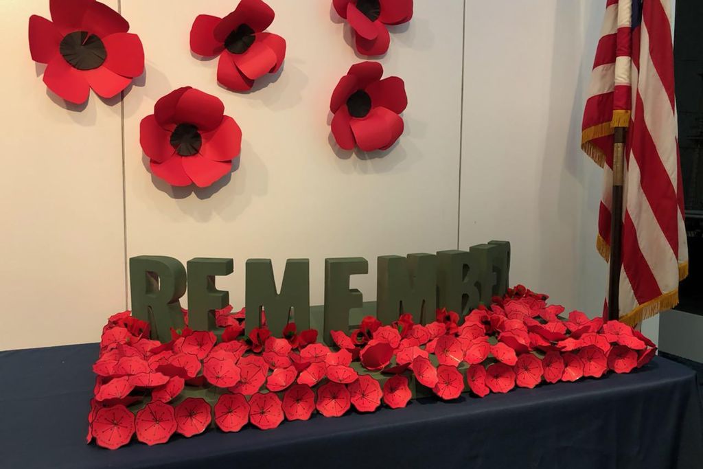 Photo of a table covered with red paper flowers around the word “Remember” in block letters. In the background, an American flag on a pole is seen.