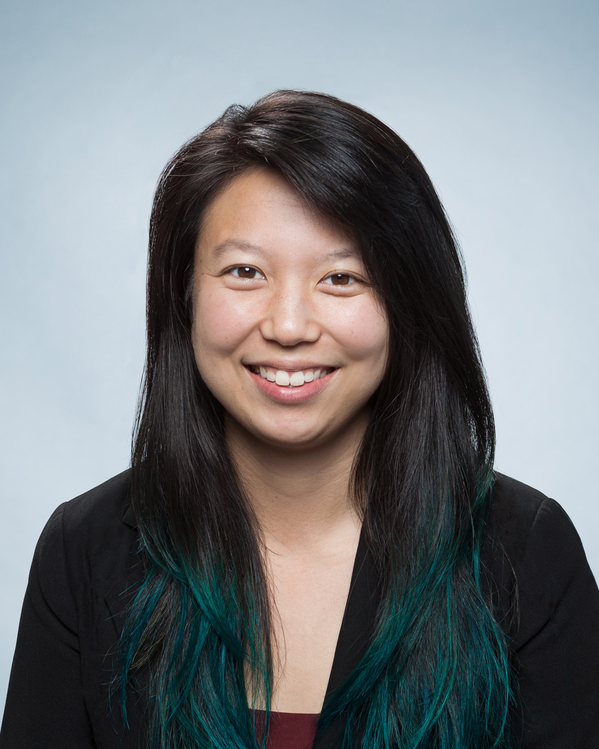 Headshot of Lena Landaverde in a black blazer smiling, in front of a light blue background. She has long black hair with some bluish teal in it.